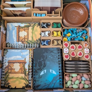 May include: A board game organizer with various components. Includes notebooks, game pieces, and bowls. The notebooks have colorful illustrations. The game pieces are in various colors and shapes. The organizer is made of light brown wood.