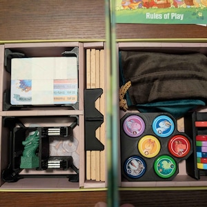 May include: Open board game box with various components. Includes cards, wooden pieces, a green dragon figurine, colorful tokens, and a drawstring bag. The box is beige with a gold clasp and the words "Rules of Play" are visible.