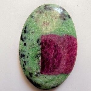 May include: Oval zoisite gemstone with a striking colour scheme. A large, rectangular patch of deep magenta-red ruby sits against a backdrop of mottled green zoisite. Black speckles add detail to the stone.