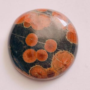 May include: A round, polished gemstone with a dark gray base and reddish-orange circular patterns. The stone has a glossy finish and a unique, natural appearance. The patterns vary in size and shape, creating a visually interesting design.