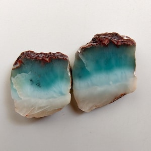 May include: Two irregular, flat slices of agate stone featuring a gradient of colors. The top edges are a rough, dark reddish-brown, transitioning into a vibrant teal blue, then fading into a translucent, milky white base with visible internal textures.