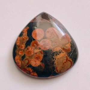 Könnte beinhalten: Ein tropfenförmiger Edelstein mit polierter Oberfläche. Der Stein hat eine dunkle Grundfarbe mit rot-orangen und braunen kreisförmigen und unregelmäßigen Mustern. Wahrscheinlich für Schmuck oder Dekorationszwecke verwendet.