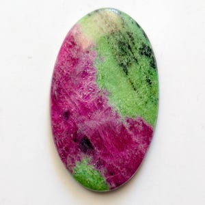 May include: Oval-shaped gemstone with a polished surface. The stone features a vibrant mix of colors, including deep magenta, bright green, and black. The magenta and green hues are the most prominent, creating a striking contrast. The gemstone is likely used for jewelry.