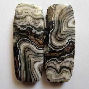 Puede incluir: Dos piedras preciosas rectangulares pulidas con un patrón arremolinado de bandas negras, blancas, marrones y grises. Las piedras tienen extremos redondeados y una superficie lisa y brillante, mostrando formaciones minerales naturales.
