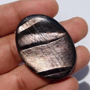 May include: An oval-shaped, polished gemstone with a dark, lustrous surface. The stone features a striking pattern of parallel, light-colored bands that create a shimmering effect. The gemstone is held in a hand.