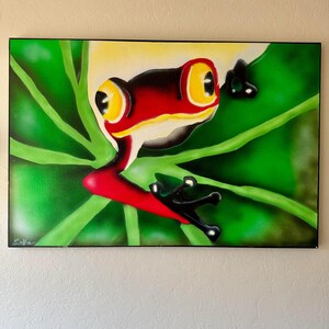 Puede incluir: Pintura enmarcada de una rana arborícola de ojos rojos. La rana tiene un cuerpo rojo, patas negras y grandes ojos amarillos. Está posada en una hoja verde con un fondo verde. La pintura está firmada en la esquina inferior izquierda.