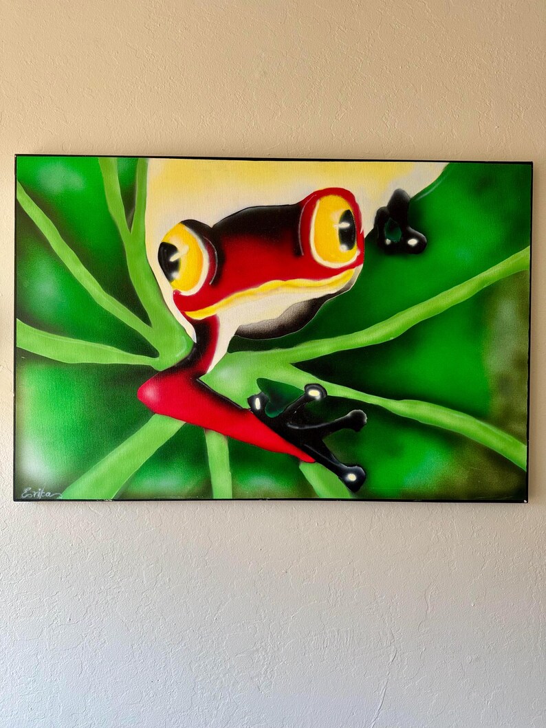 Op de afbeelding: Een ingelijst schilderij van een roodoogboomkikker. De kikker heeft een rood lichaam, zwarte poten en grote gele ogen. Hij zit op een groen blad met een groene achtergrond. Het schilderij is linksonder gesigneerd.