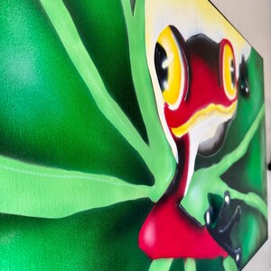Op de afbeelding: Een levendig schilderij van een rood-zwarte kikker met gele ogen, tegen een achtergrond van weelderige groene bladeren. Het kunstwerk staat op canvas en toont een kleurrijke en gedetailleerde weergave van de amfibie in zijn natuurlijke habitat.