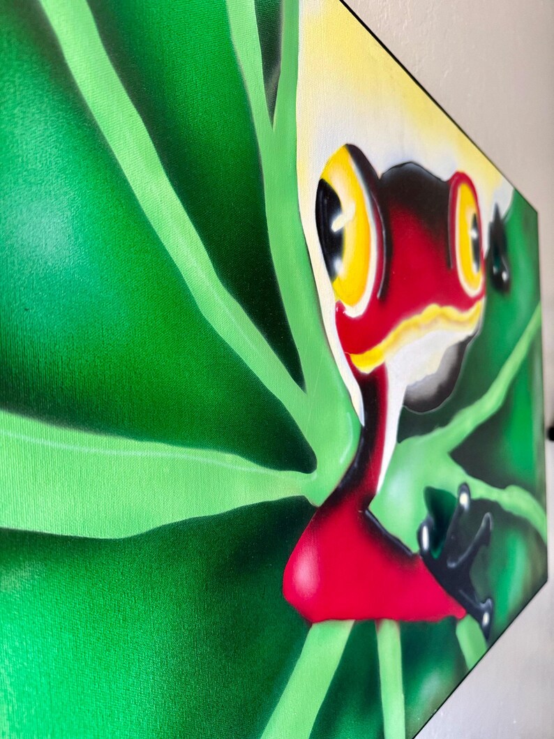 Op de afbeelding: Een levendig schilderij van een rood-zwarte kikker met gele ogen, tegen een achtergrond van weelderige groene bladeren. Het kunstwerk staat op canvas en toont een kleurrijke en gedetailleerde weergave van de amfibie in zijn natuurlijke habitat.
