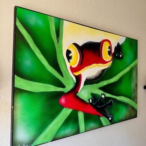 Op de afbeelding: Een ingelijst schilderij van een roodoogboomkikker op een groen blad. De kikker heeft een rode kop, gele ogen en zwarte poten. De achtergrond is een verloop van groen. De handtekening van de kunstenaar, "Erika", staat linksonder.