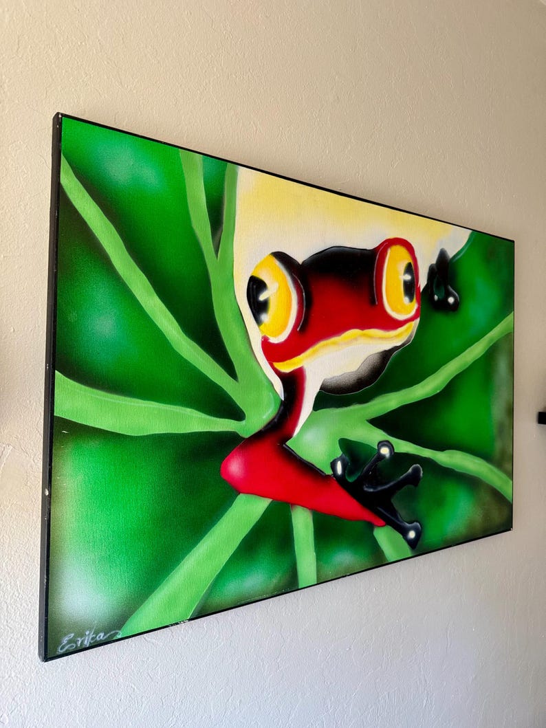 Op de afbeelding: Een ingelijst schilderij van een roodoogboomkikker op een groen blad. De kikker heeft een rode kop, gele ogen en zwarte poten. De achtergrond is een verloop van groen. De handtekening van de kunstenaar, "Erika", staat linksonder.