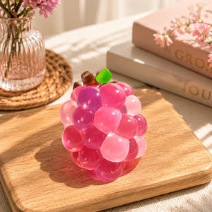 Peut inclure: Un amas de perles de gel roses et magenta, ressemblant à une grappe de raisin, est posé sur une planche à découper en bois. Les perles ont une tige verte et marron. Un petit vase de fleurs roses et un livre sont en arrière-plan.