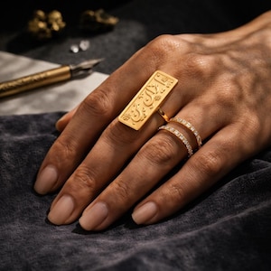 Peut inclure: Une bague dorée avec un motif rectangulaire gravé et des caractères arabes. La bague est portée sur un doigt, associée à une bague à double rangée ornée de petites pierres scintillantes. Le bijou est sur fond sombre.