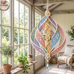 May include: A hanging crochet ornament featuring pastel pink, blue, yellow, and purple lattice-patterned ribbons arranged in a spherical shape. The piece is accented with three white daisy flowers with yellow centers and a clear crystal drop.