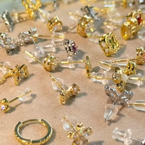 Könnte beinhalten: Eine Auswahl an goldenen und silbernen Ohrsteckern mit verschiedenen Designs, darunter Blumen-, geometrische und Edelsteinakzente. Die Ohrringe werden auf einer neutralen Oberfläche präsentiert, die ihre komplizierten Details und funkelnden Verzierungen hervorhebt. Ein goldener Creolenohrring ist ebenfalls sichtbar.