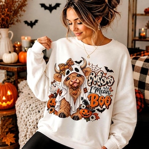 Puede incluir: Una sudadera blanca de manga larga con un gráfico de una vaca Highland disfrazada de fantasma con un lazo de leopardo. El texto dice Moo I Mean Boo, rodeado de calabazas, murciélagos, una telaraña y hojas de otoño en naranja y negro.