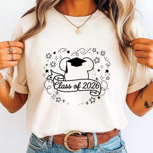 May include: Cream-colored t-shirt featuring a black graphic design. The design includes a chef's hat with a graduation cap, a banner with the text "Class of 2026", and star accents. The shirt is worn with blue jeans and a brown belt.