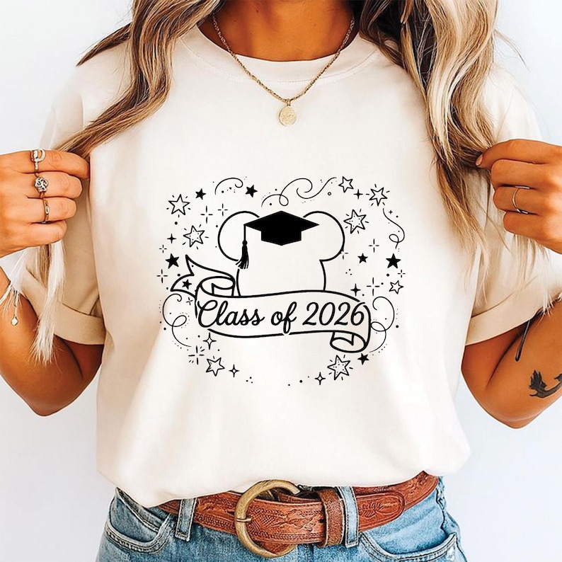 May include: Cream-colored t-shirt featuring a black graphic design. The design includes a chef's hat with a graduation cap, a banner with the text "Class of 2026", and star accents. The shirt is worn with blue jeans and a brown belt.