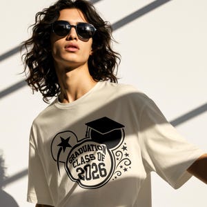 May include: Cream-colored t-shirt with a black graduation-themed graphic. The design includes a graduation cap, stars, and the text "GRADUATION CLASS OF 2026". The person in the image is wearing sunglasses.