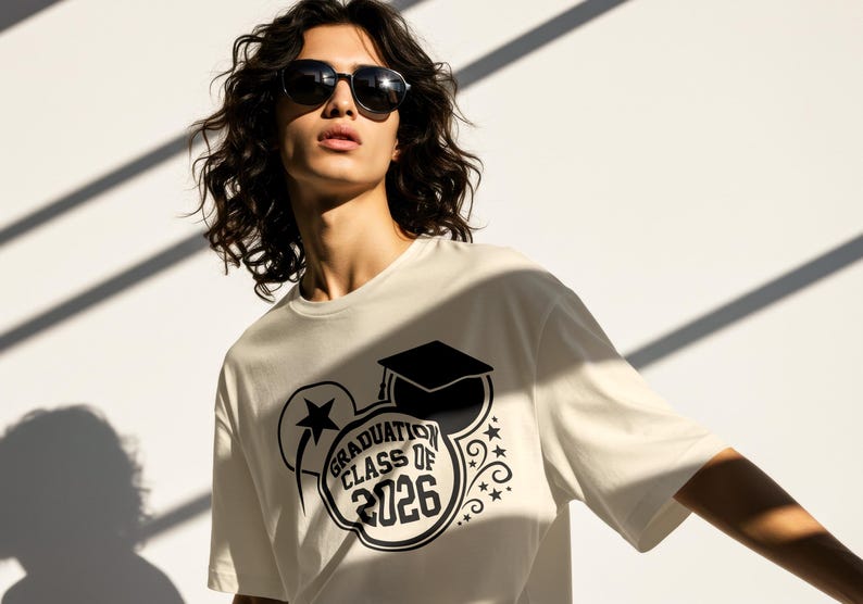 May include: Cream-colored t-shirt with a black graduation-themed graphic. The design includes a graduation cap, stars, and the text "GRADUATION CLASS OF 2026". The person in the image is wearing sunglasses.