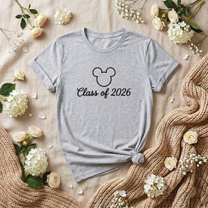 May include: A heather gray t-shirt featuring a black Mickey Mouse head outline and the words "Class of 2026" in a script font. The shirt is displayed on a neutral-toned surface with floral accents.