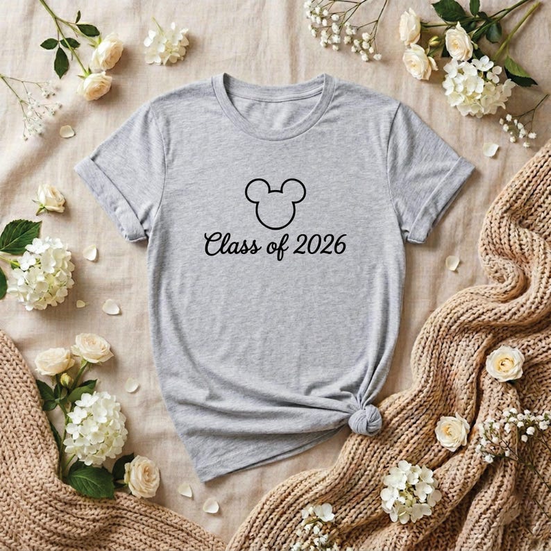 May include: A heather gray t-shirt featuring a black Mickey Mouse head outline and the words "Class of 2026" in a script font. The shirt is displayed on a neutral-toned surface with floral accents.