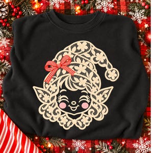 May include: A black crewneck sweatshirt featuring a graphic of a smiling elf face wearing a hat made of cream-colored lace with floral patterns. The elf has pink cheeks and a small red and white checkered bow attached to the side of the hat.