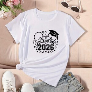 May include: White t-shirt with a black graphic that says "Class of 2026" with a graduation cap and Mickey Mouse ears design. The shirt is paired with denim shorts and white sneakers. Sunglasses are on the top right.