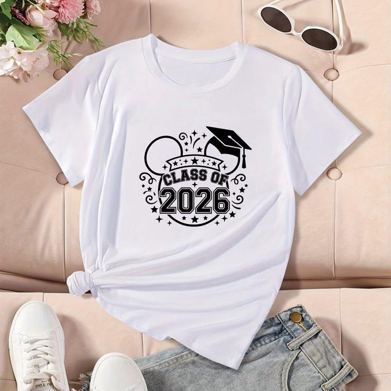 May include: White t-shirt with a black graphic that says "Class of 2026" with a graduation cap and Mickey Mouse ears design. The shirt is paired with denim shorts and white sneakers. Sunglasses are on the top right.