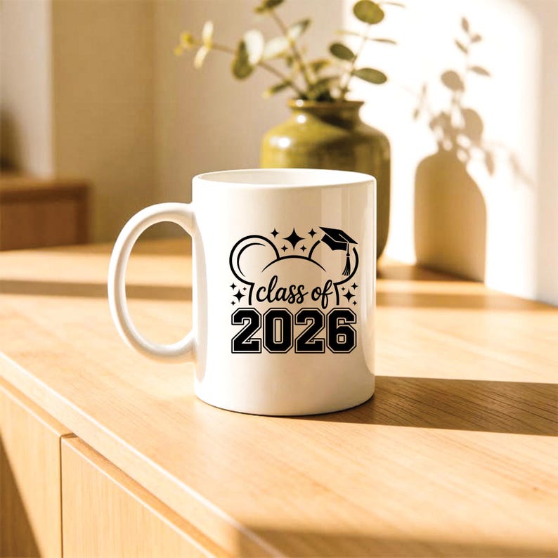 May include: White ceramic mug with a black graphic that reads "Class of 2026". The design includes a graduation cap, stars, and Mickey Mouse ears. The mug is on a wooden surface.