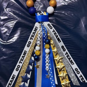 May include: A spirit mum accessory featuring a circular ring of white, navy blue, and gold beads. Multiple ribbons hang down, including white ribbons with the text GO TEAM!, gold glitter ribbons, navy blue ribbons with star cutouts, and a central chain with beads and the text LOPEZ LOBOS.