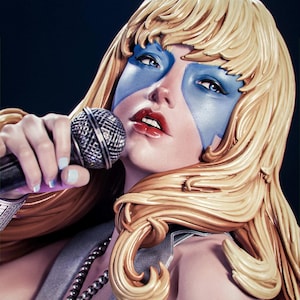 Puede incluir: Escultura de una persona con cabello rubio, pintura facial azul y lápiz labial rojo sosteniendo un micrófono. La persona lleva un collar plateado con un colgante de bola de discoteca. El fondo es oscuro.