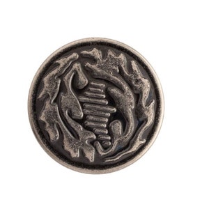 May include: A round, antique silver button with a detailed design. The button features a central, stacked rectangular shape, surrounded by stylized, flowing organic forms. The button has a raised, textured surface and a dark, aged appearance.