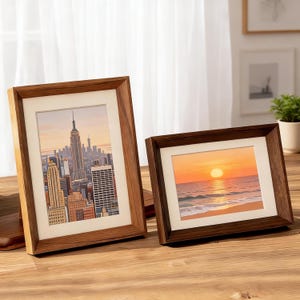 Op de afbeelding: Twee houten fotolijsten. Eén lijst toont een stadsgezicht met het Empire State Building, de andere een zonsondergang boven de oceaan. De lijsten hebben een lichtbruine en donkerbruine afwerking, met een witte passe-partout rond de afbeeldingen.