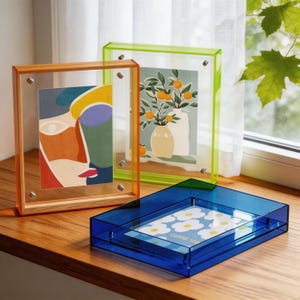 May include: Three acrylic picture frames in orange, green, and blue. The orange frame has a wooden border and holds an abstract face art print. The green frame displays a vase of oranges. The blue frame holds a floral print.