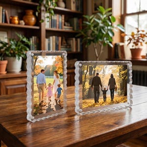 Op de afbeelding: Twee heldere acryl fotolijsten met golvende randen. Elke lijst bevat een kleurenfoto van een gezin dat buiten wandelt. De lijsten staan op een donkere houten tafel in een goed verlichte kamer.