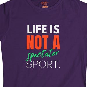 May include: A purple t-shirt with the text "LIFE IS NOT A spectator SPORT." in white, orange, and green. The shirt has a crew neck and short sleeves. The brand logo is at the top.