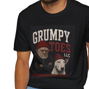 May include: Black t-shirt featuring a graphic of an elderly man and a dog, both wearing red beanies. The text "GRUMPY TOES LLC" is above the image in white and red.