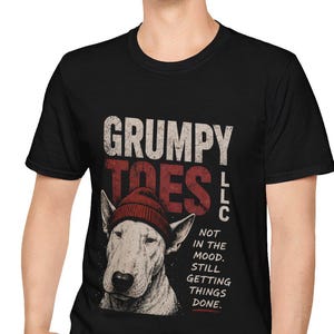 May include: Black t-shirt with the text "GRUMPY TOES LLC" in white and red. The shirt features a graphic of a Bull Terrier wearing a red beanie. The text "NOT IN THE MOOD. STILL GETTING THINGS DONE." is also printed on the shirt.