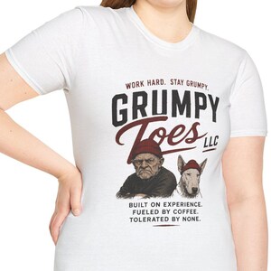 May include: White t-shirt with the text "GRUMPY TOES LLC" and illustrations of a grumpy man and a dog. The shirt also includes the phrases "WORK HARD. STAY GRUMPY" and "BUILT ON EXPERIENCE. FUELED BY COFFEE. TOLERATED BY NONE."