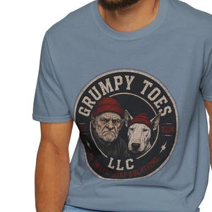 May include: A light blue t-shirt featuring a circular graphic with the text "GRUMPY TOES" and the year "2024". The graphic includes illustrations of an elderly man and a dog wearing red hats. The shirt is made of a soft material.