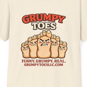 May include: A cream-colored short-sleeve t-shirt featuring a graphic of five cartoon toes with angry facial expressions. The text reads GRUMPY TOES, FUNNY. GRUMPY. REAL., and GRUMPYTOESLLC.COM. The shirt has a speckled texture throughout the fabric.