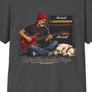 May include: Dark gray t-shirt featuring a graphic of a man playing a red guitar, with a dog and Marshall amplifiers. The text "Enjoy the simple things" is below the illustration. The shirt has a crew neck and short sleeves.