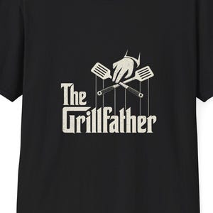 Puede incluir: Camiseta negra con un gráfico que dice "The Grillfather". El gráfico presenta una mano sosteniendo utensilios de parrilla cruzados, diseñado al estilo de la película "El Padrino".