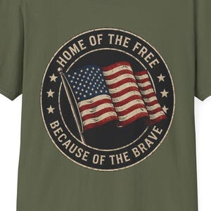 May include: Olive green t-shirt featuring a distressed graphic of the American flag within a circular emblem. The emblem includes the text "HOME OF THE FREE" and "BECAUSE OF THE BRAVE".