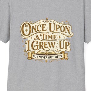 May include: A heather gray t-shirt featuring the text "Once Upon a Time I Grew Up But Never Out of It" in a vintage-style design. The text is in gold and brown tones, with decorative flourishes and a book illustration.
