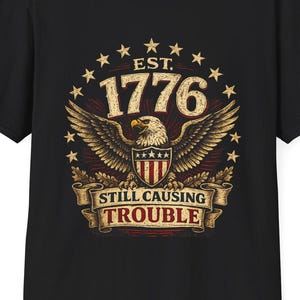 May include: A black short-sleeve t-shirt featuring a graphic of a bald eagle with wings spread, a shield with red and white stripes, and stars. The text reads EST. 1776 and STILL CAUSING TROUBLE in a vintage-style font with a beige and red color palette.