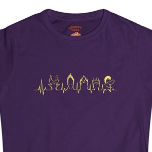 May include: A purple t-shirt featuring a gold heartbeat design with silhouettes of iconic villains. The shirt has a crew neck and short sleeves. The text "GRUMPY LOVE" is printed on the upper right side of the shirt.