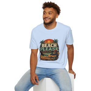 A smiling man wearing a white t-shirt with a graphic design of a beach scene and the text "BEACH PLEASE" is sitting on a white stool.