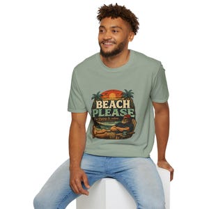 A smiling man wearing a green t-shirt with a graphic design of a beach scene and the text "BEACH PLEASE" is sitting on a white stool.